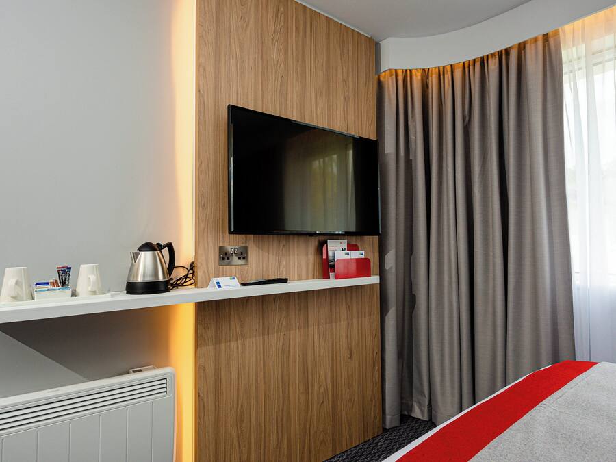 A hotel room with a flat-screen TV mounted on a wooden wall, a white shelf with a kettle, a remote control, and a magazine rack.