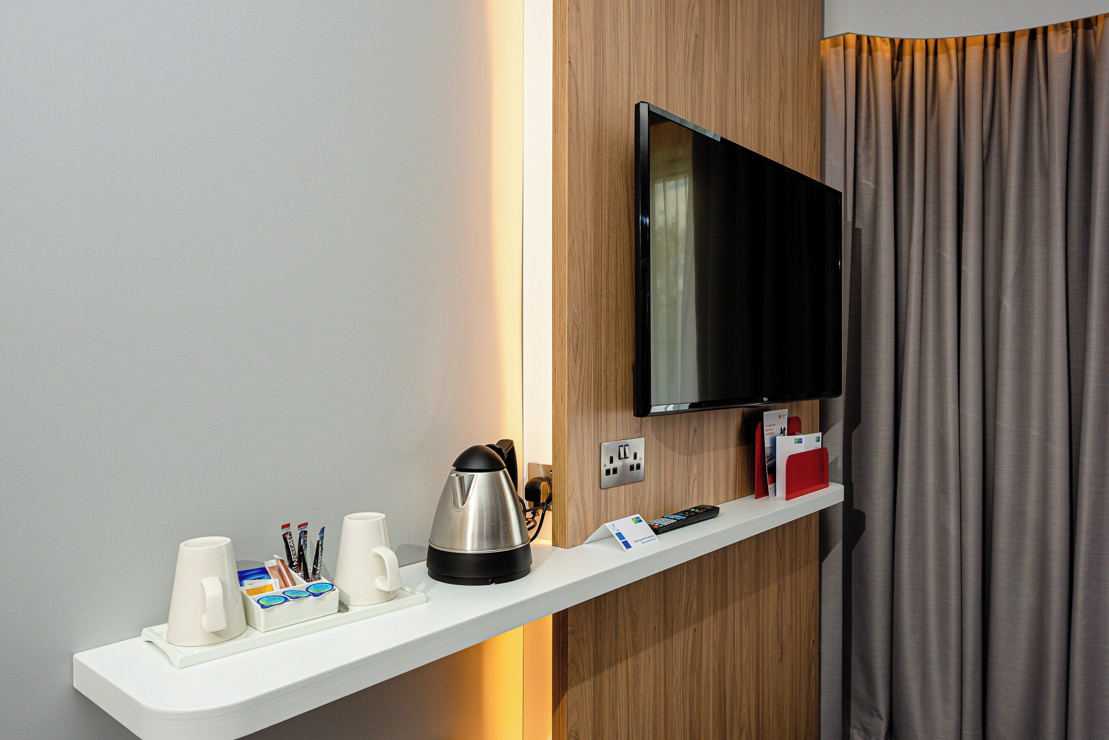 A hotel room with a flat-screen TV mounted on a wooden panel, a white shelf with a kettle, cups, and a remote control.