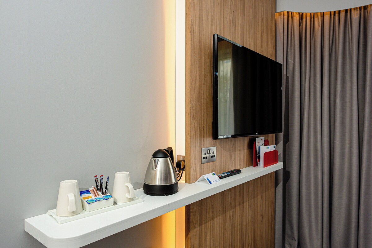 Habitación de hotel con televisión de pantalla plana fijada a un panel de madera, repisa blanca con tetera, tazas y control remoto.