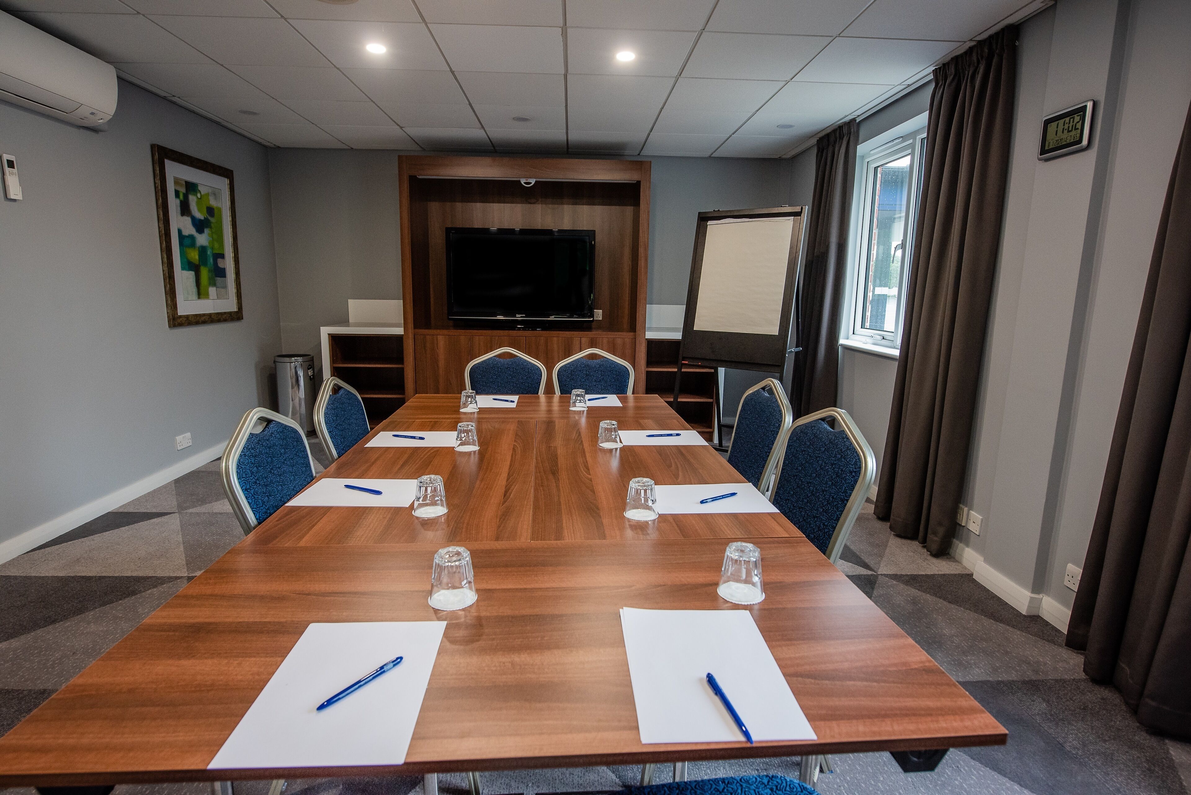 A conference room with a long wooden table, blue chairs, a projector screen, and a large flat-screen TV.