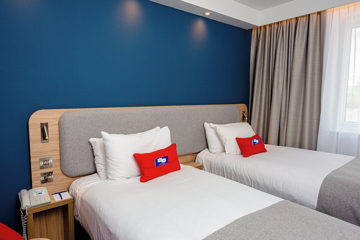 Habitación de hotel con dos camas, cabecero de madera, mesita de noche con teléfono y bloc de notas, y ventana con cortinas.