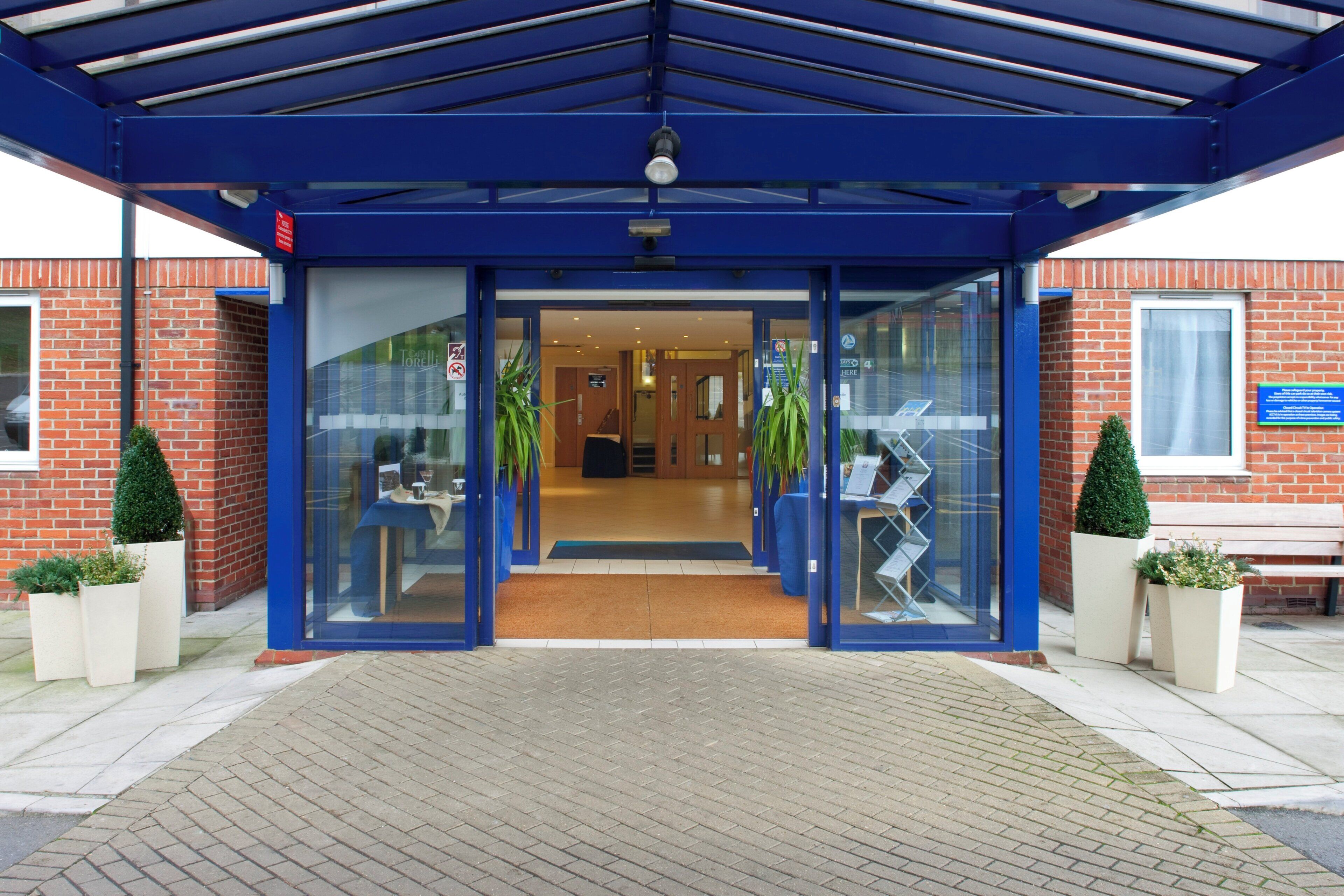 Entrada de hotel con toldo azul, puertas de vidrio y plantas en macetas.