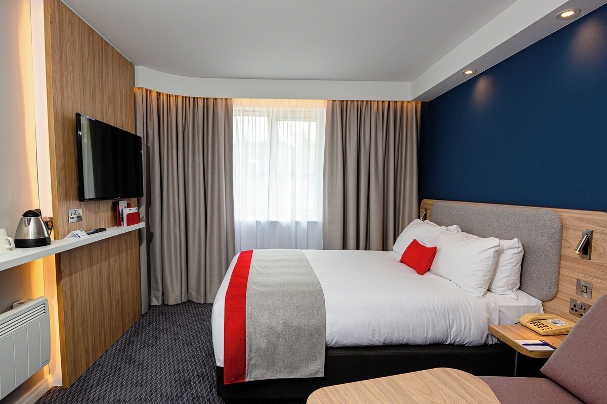 Una habitación de hotel con cama, televisión, escritorio con teléfono y ventana con cortinas.