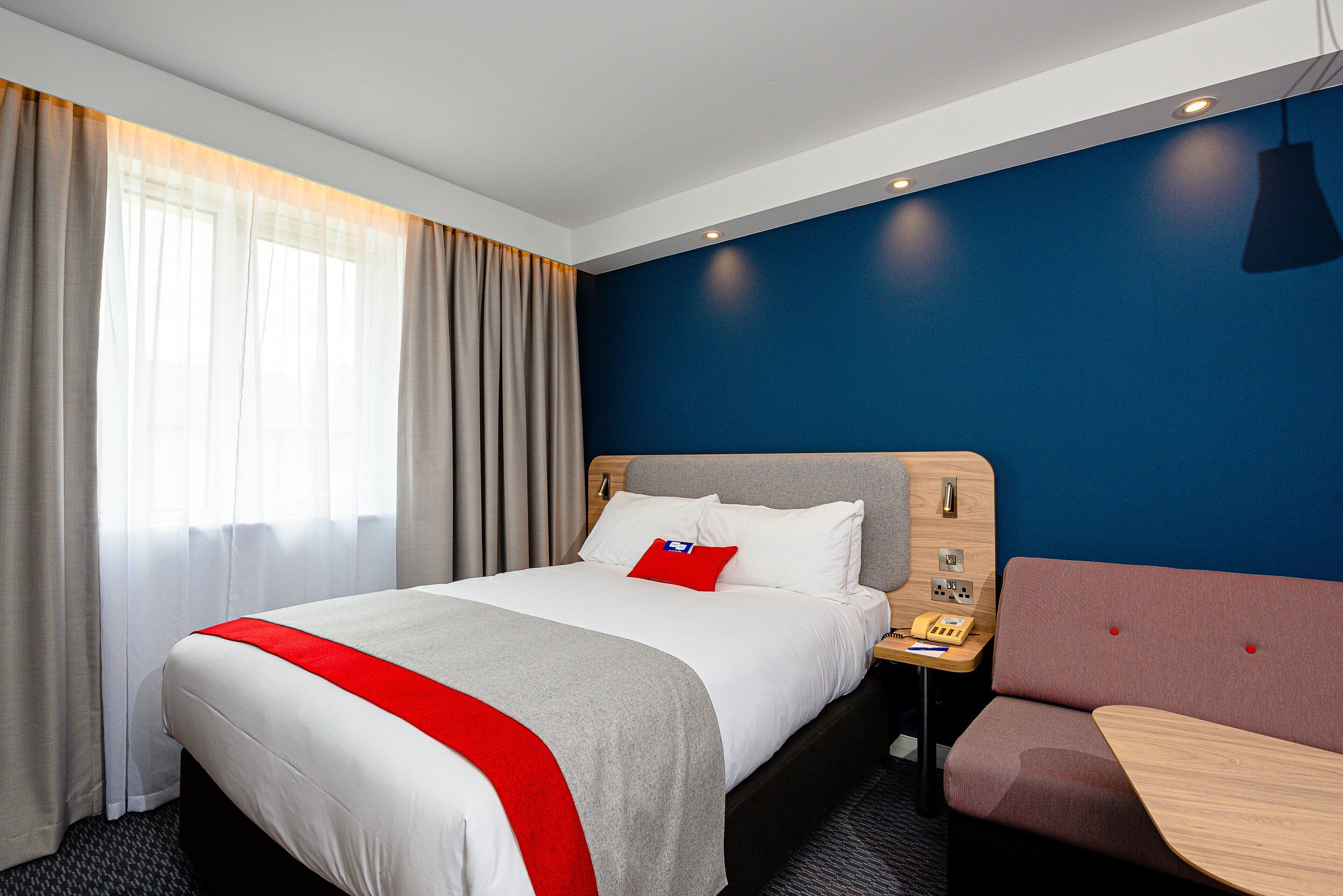 A hotel room with a bed, a bedside table with a phone, a red pillow, and a seating area with a table.