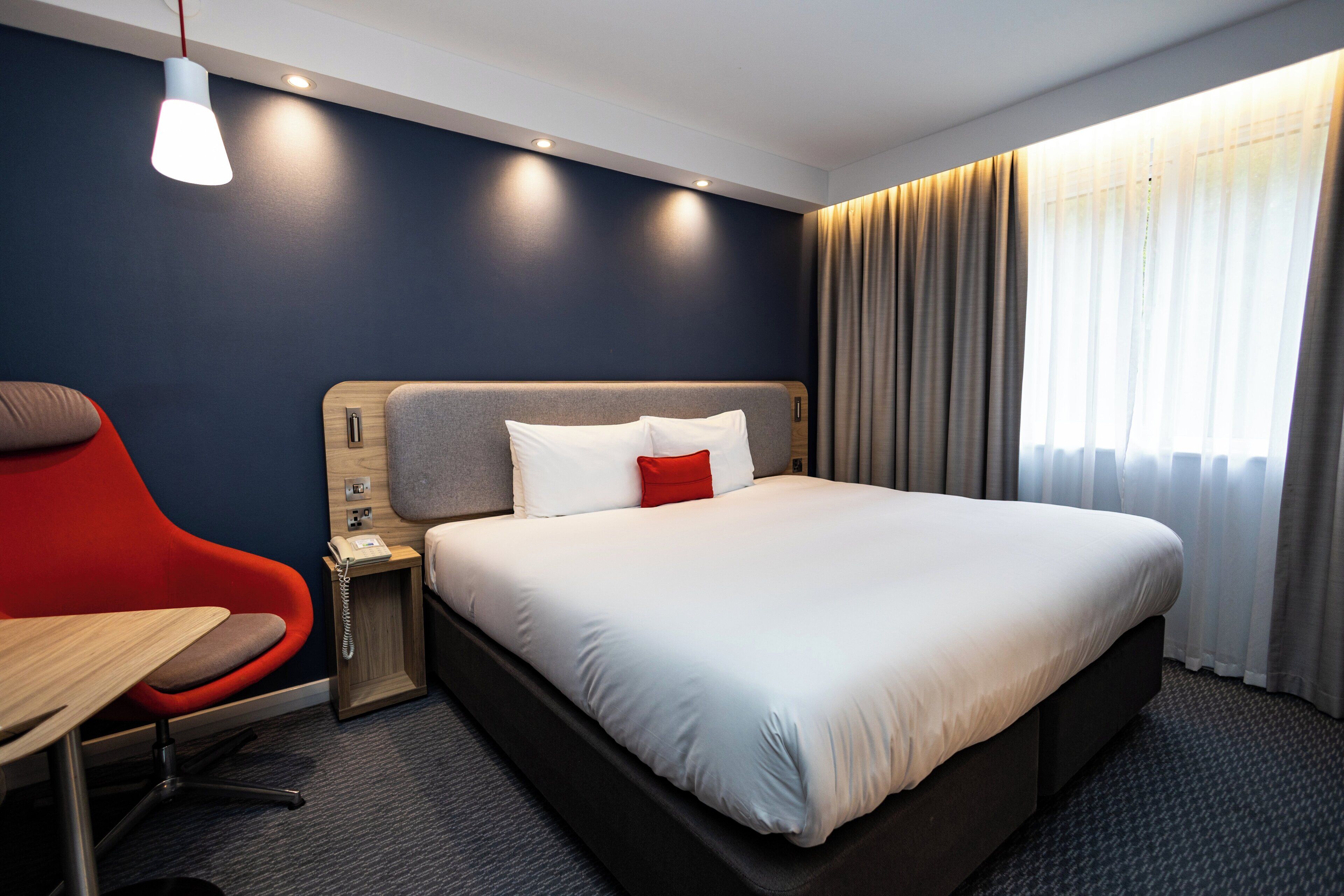 Habitación de hotel con cama, escritorio, silla, mesita de noche y ventana con cortinas.