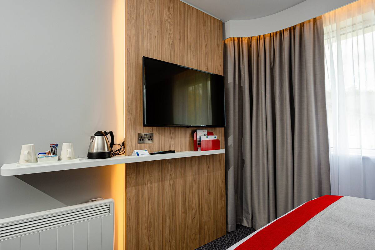A hotel room with a flat-screen TV mounted on a wooden wall, a white shelf with a kettle and cups, a red and grey bedspread, and a large window with grey curtains.