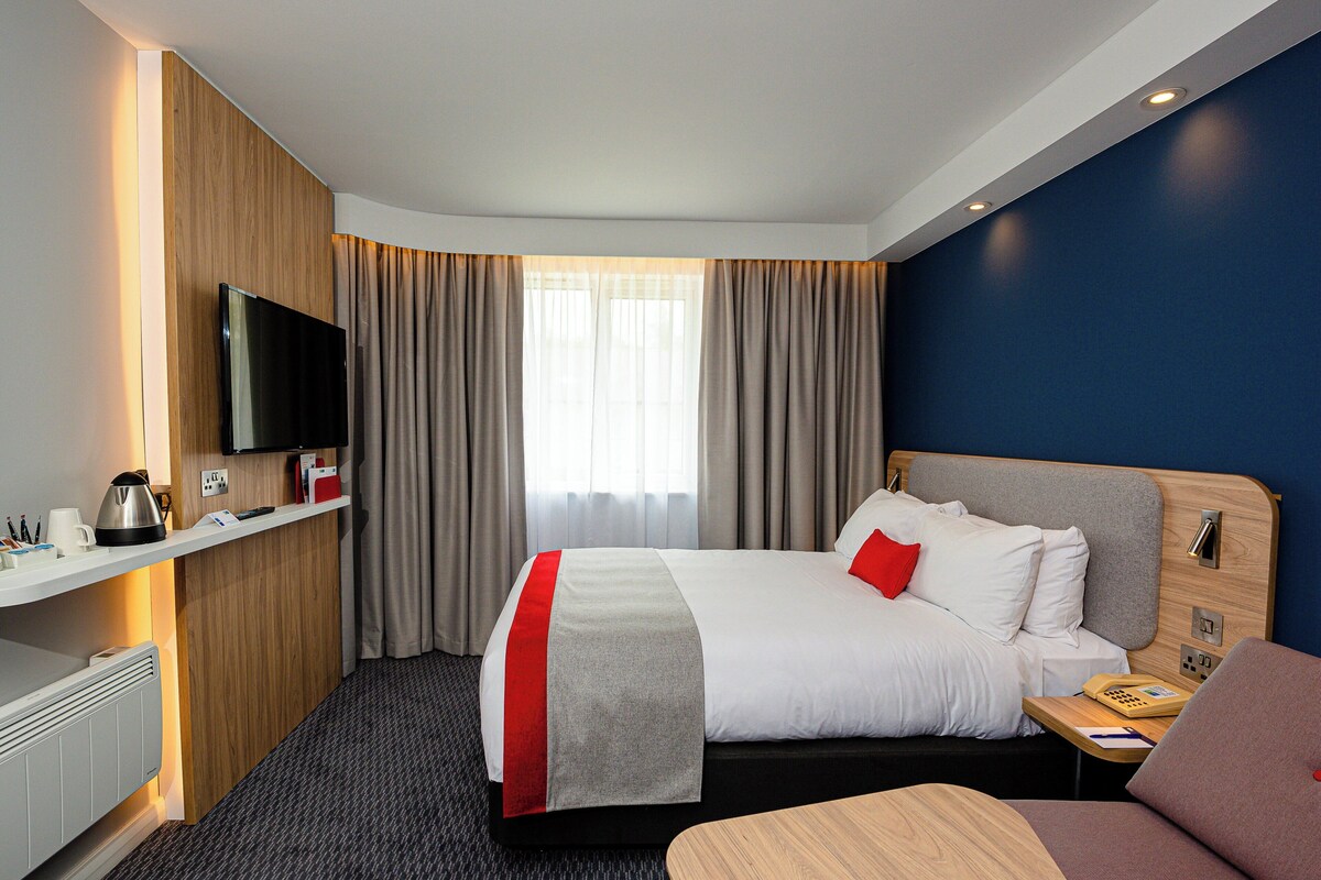 Habitación de hotel con cama, escritorio, televisor de pantalla plana, teléfono y ventana con cortinas.