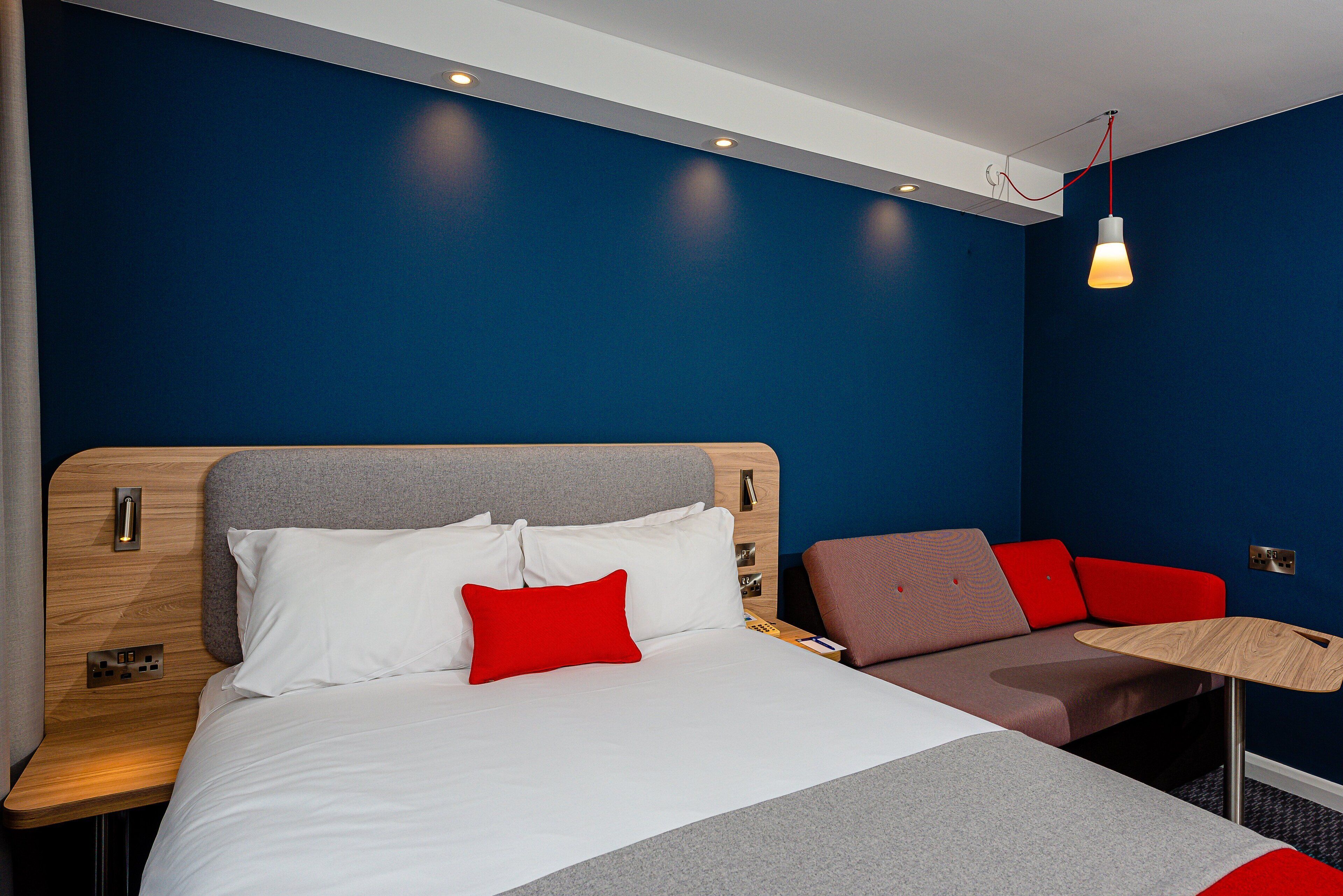 A modern hotel room with a large bed, a bedside table, a desk, and a red and brown sofa.