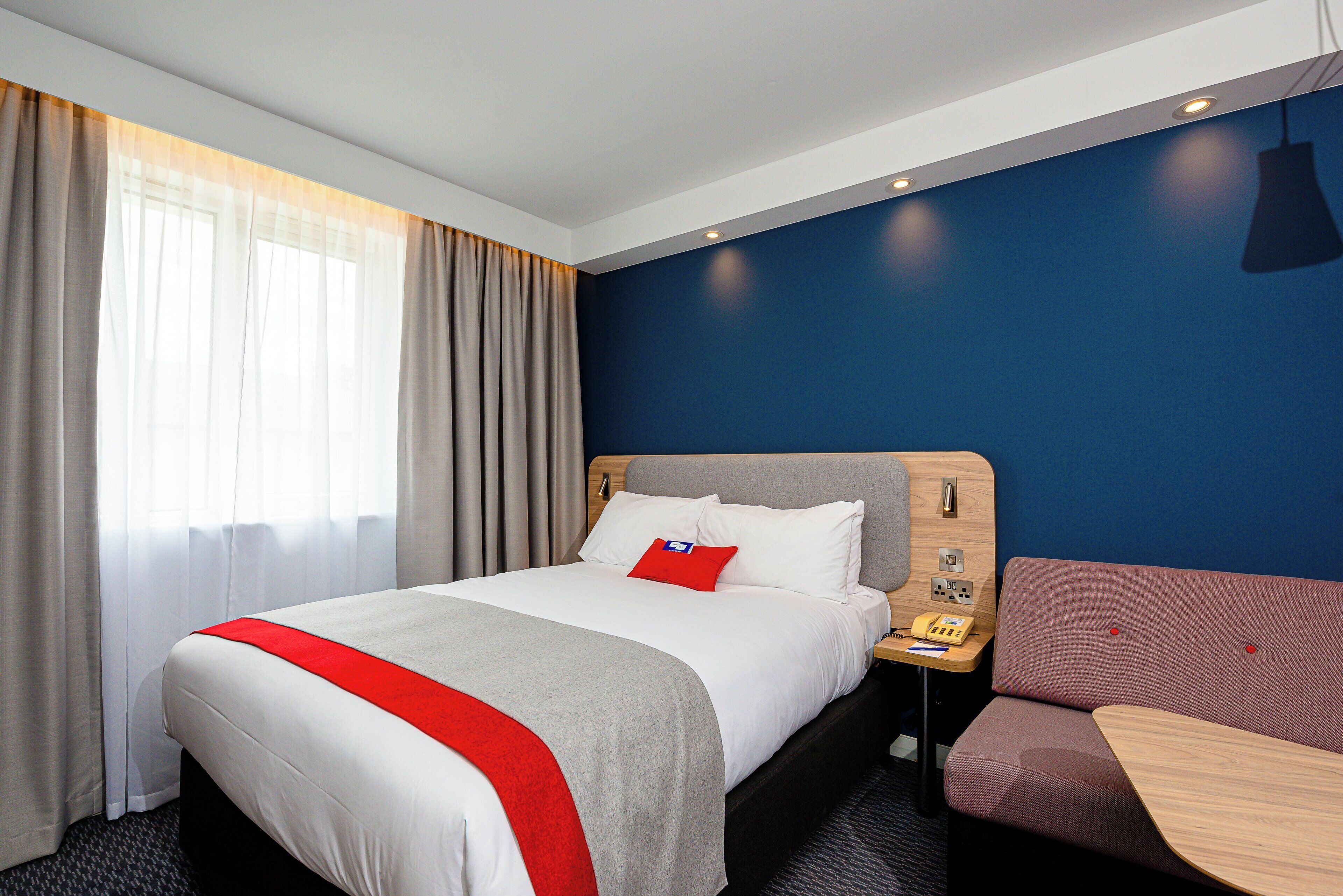 Ein Hotelzimmer mit einem Bett, einem Nachttisch, einem Telefon, einem roten Kopfkissen, einer Couch und einem Fenster mit Vorhängen.