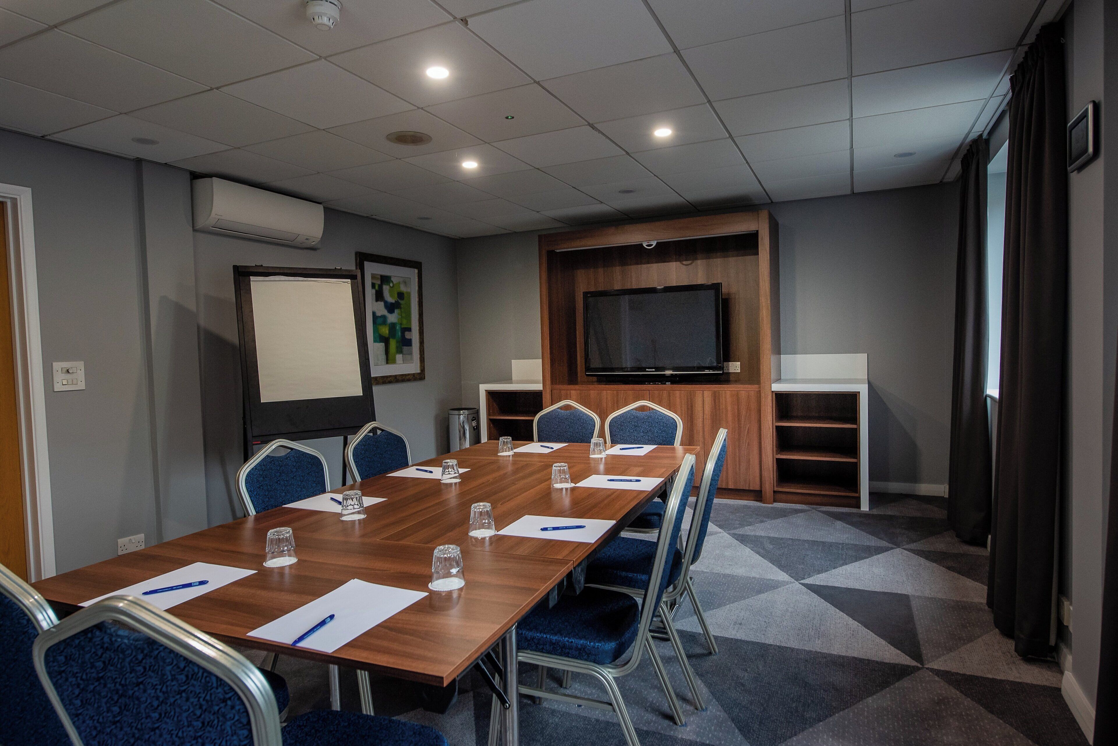 A conference room with a long table, blue chairs, a projector screen, a television, and a small shelf.