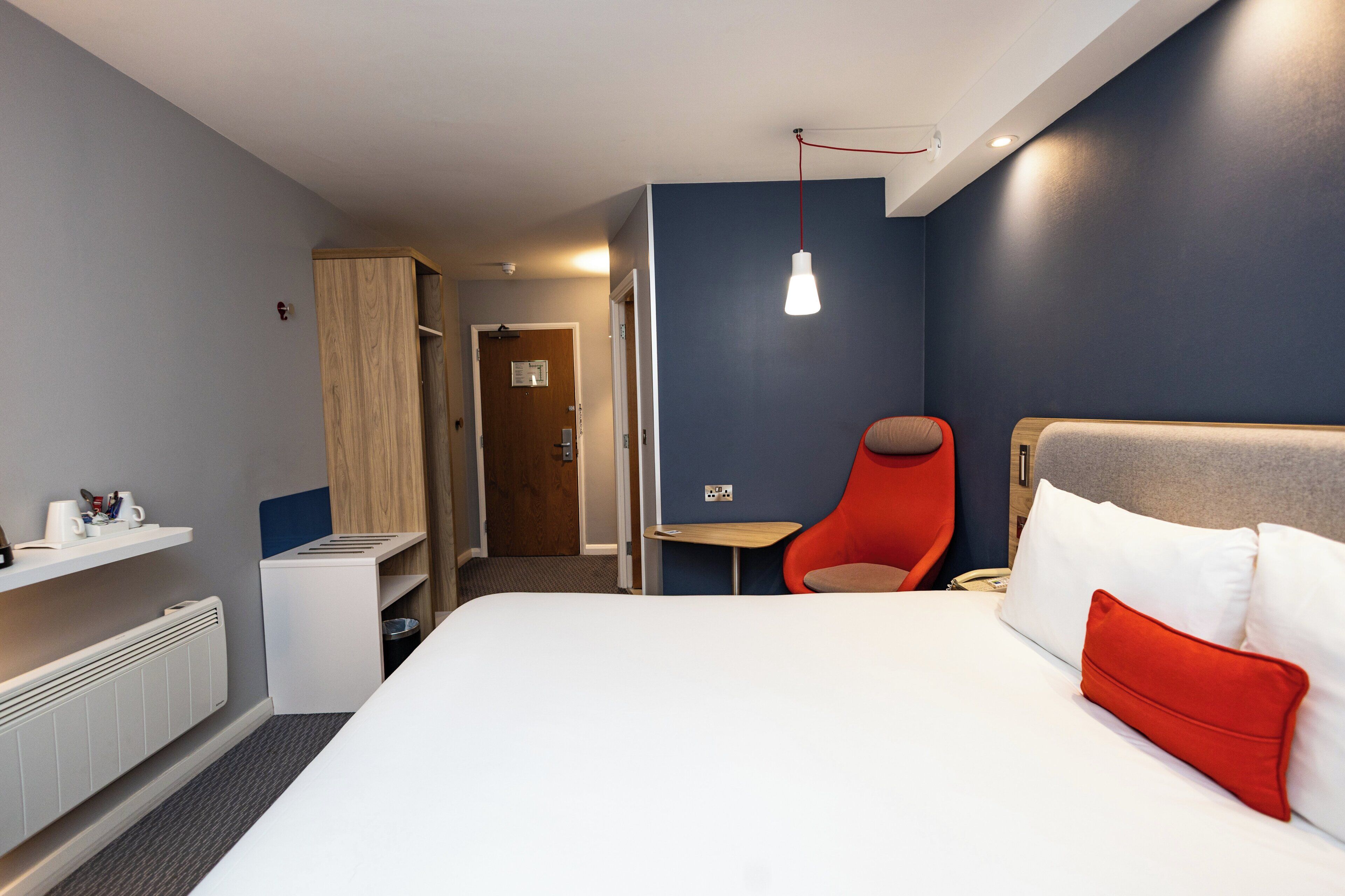 A hotel room with a bed, a chair, a small table, a heater, and a door leading to another room.