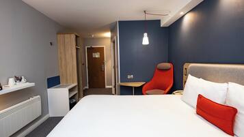 A hotel room with a bed, a chair, a small table, a heater, and a door leading to another room.
