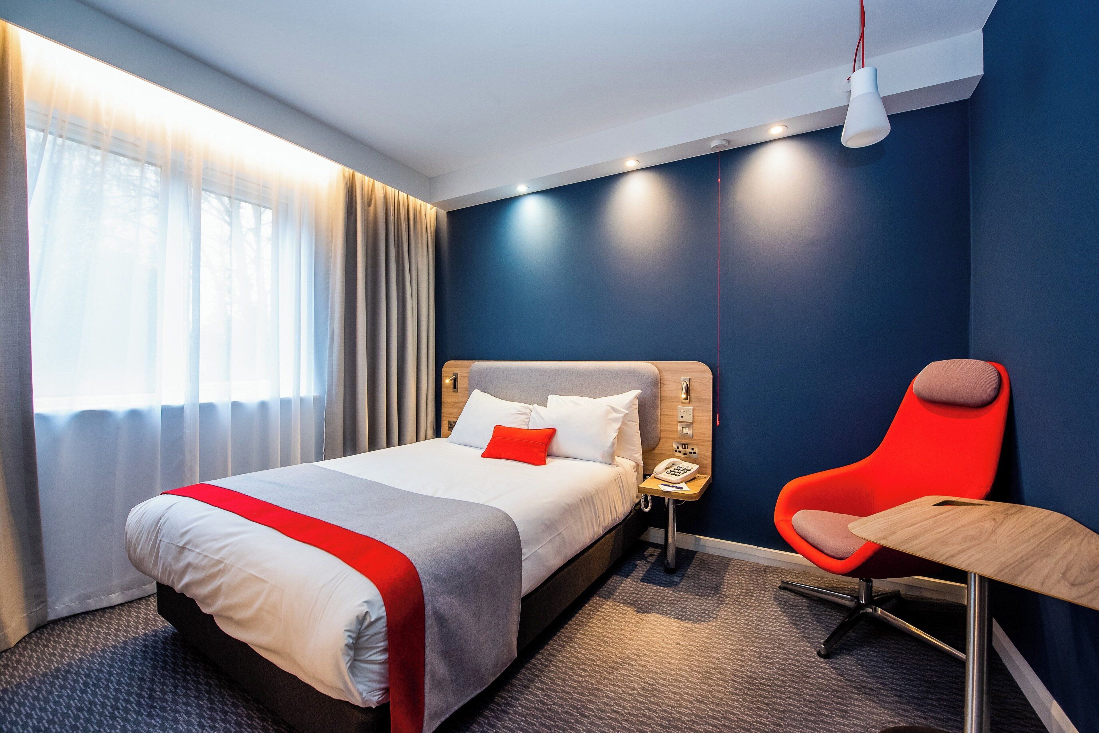 Una habitación de hotel con una cama, una silla roja, una mesita y un teléfono.