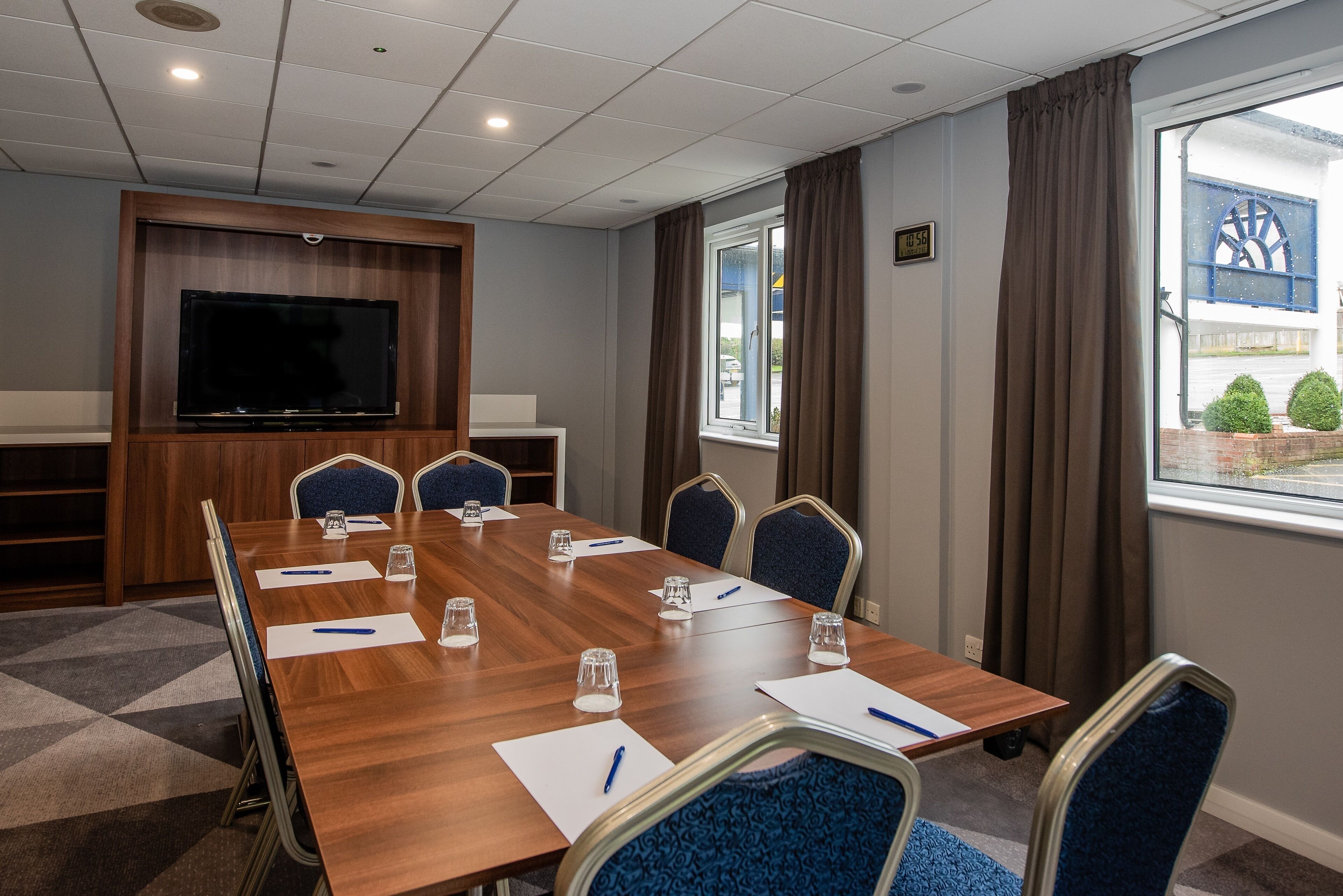 A conference room with a long table, chairs, a TV, and a window with curtains.