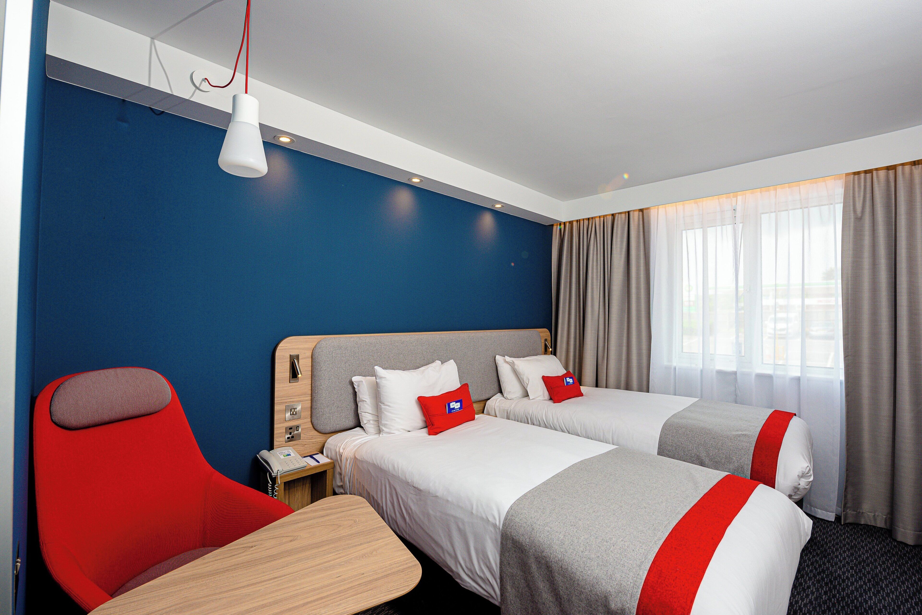 Habitación de hotel con dos camas, una silla roja, una mesa de madera y una ventana con cortinas.