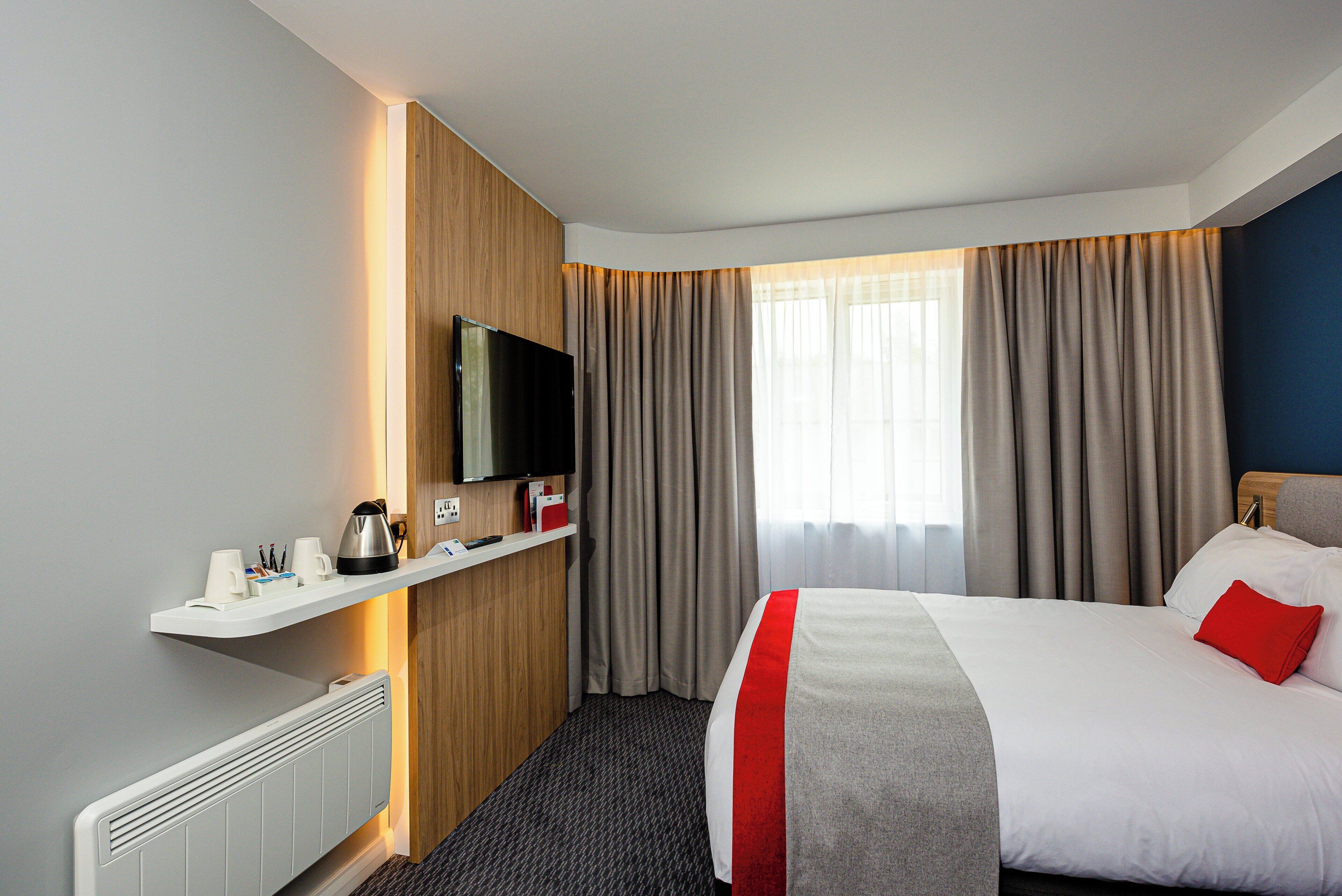 Habitación de hotel con cama, televisor, repisa con artículos de aseo y ventana con cortinas.