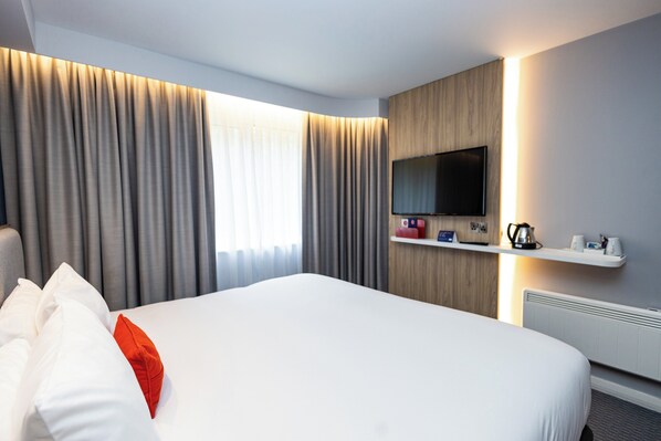 Una habitación de hotel con una cama grande, cortinas, un televisor montado y una estantería con objetos.