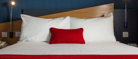 Una cama bien hecha con sábanas blancas y un cojín rojo decorativo, con dos lámparas de noche a cada lado.
