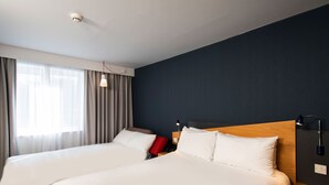 Une chambre d’hôtel avec deux lits, une fenêtre avec des rideaux, une table de chevet avec une lampe et un luminaire fixé au mur.