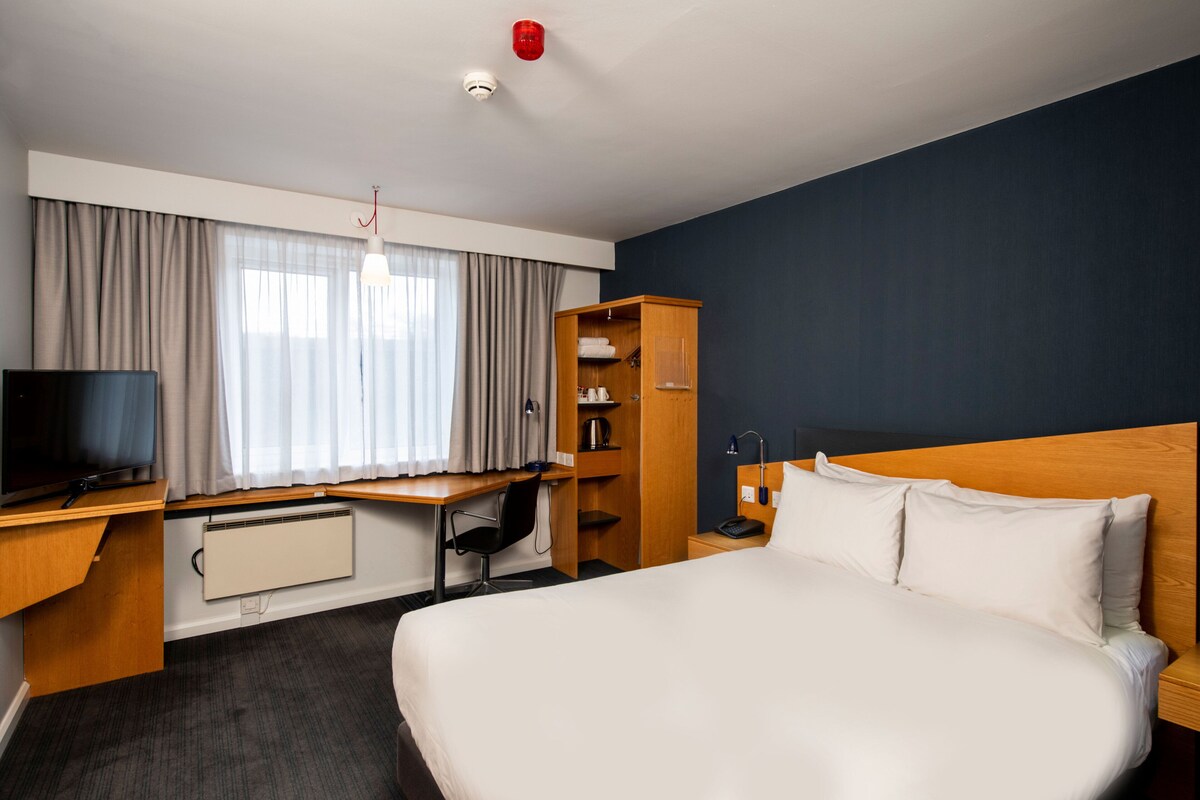 A hotel room with a large bed, a desk with a computer, a television, a wardrobe, and a window with curtains.