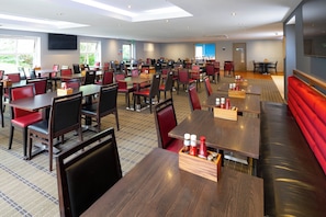 A spacious dining area with multiple tables and chairs, a flat-screen TV mounted on the wall, and a view of the outdoors through large windows. - Holiday Inn Express Exeter East by IHG (Exeter)