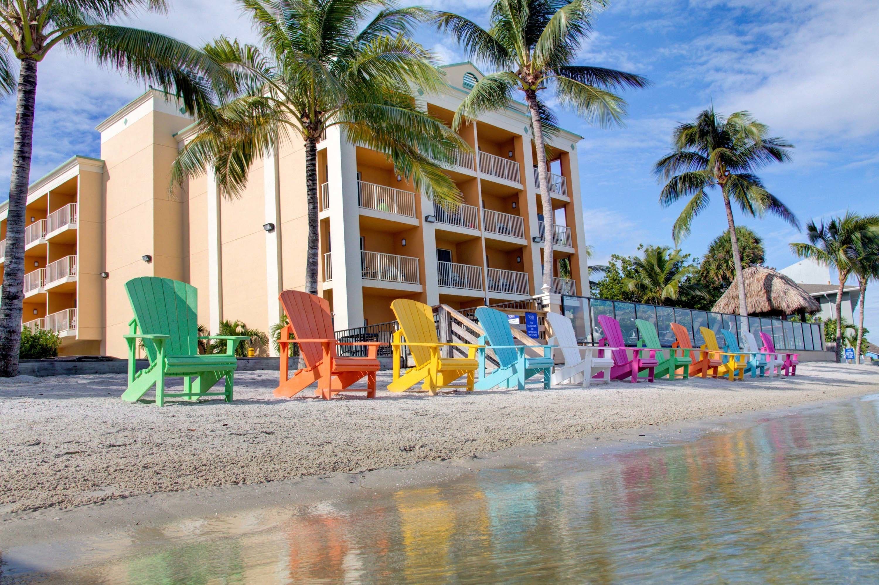 A hotel with multiple balconies, a beachside location, and colorful Adirondack chairs lined up on the sand.