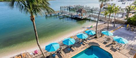 A pool area with lounge chairs and umbrellas, a beach with a thatched roof structure, and a pier with boats.