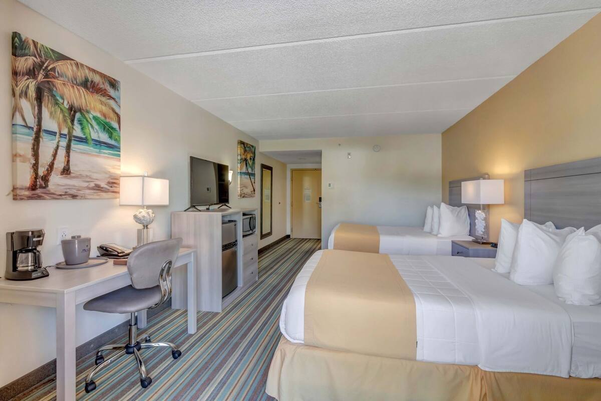 A hotel room with two beds, a desk, a chair, a TV, and a painting of palm trees.
