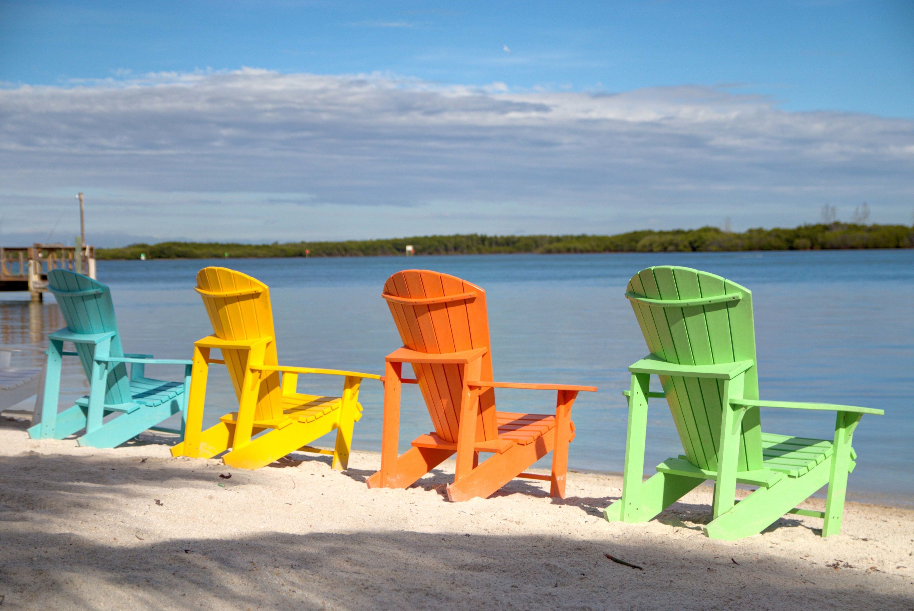 A row of colorful Adirondack chairs on a sandy beach with a calm body of water and a distant shoreline in the background.