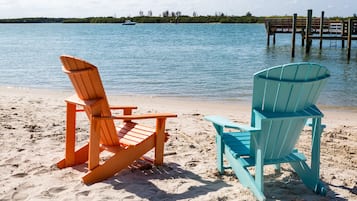 Two beach chairs on a sandy shore with a body of water and a dock in the background.
