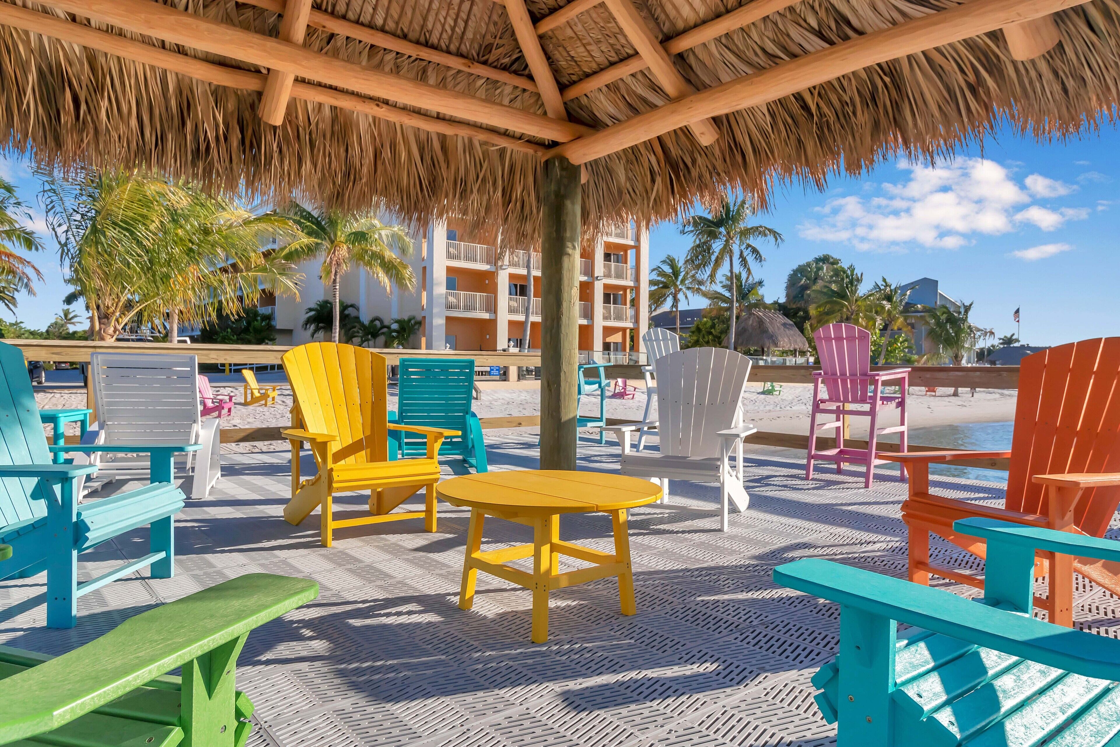 Un espacio de descanso frente a la playa con sillas y mesas tipo Adirondack de colores, techo de paja y vistas a un hotel y palmeras.