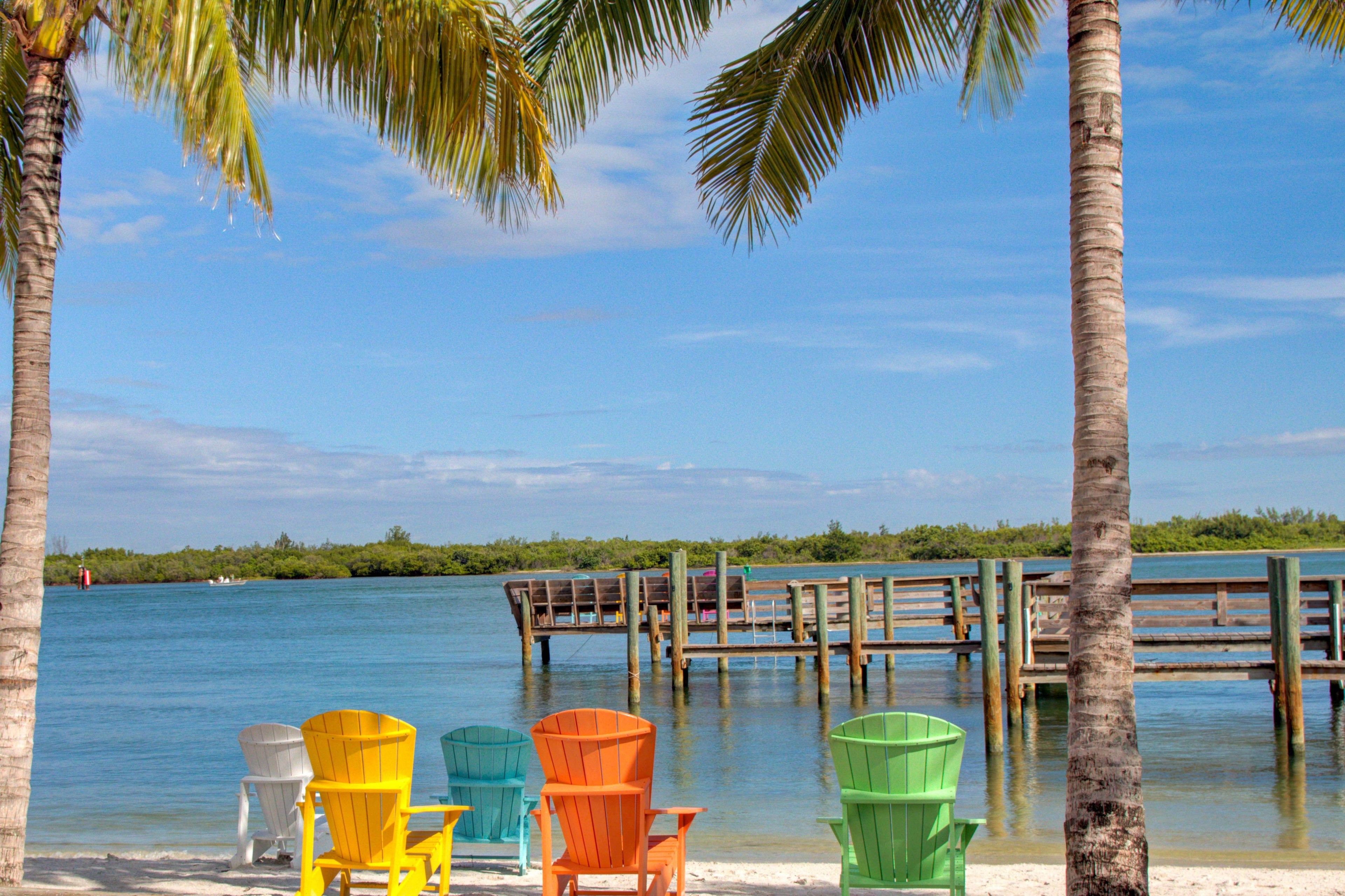 A beach with colorful Adirondack chairs, a wooden dock, and a clear blue sky.