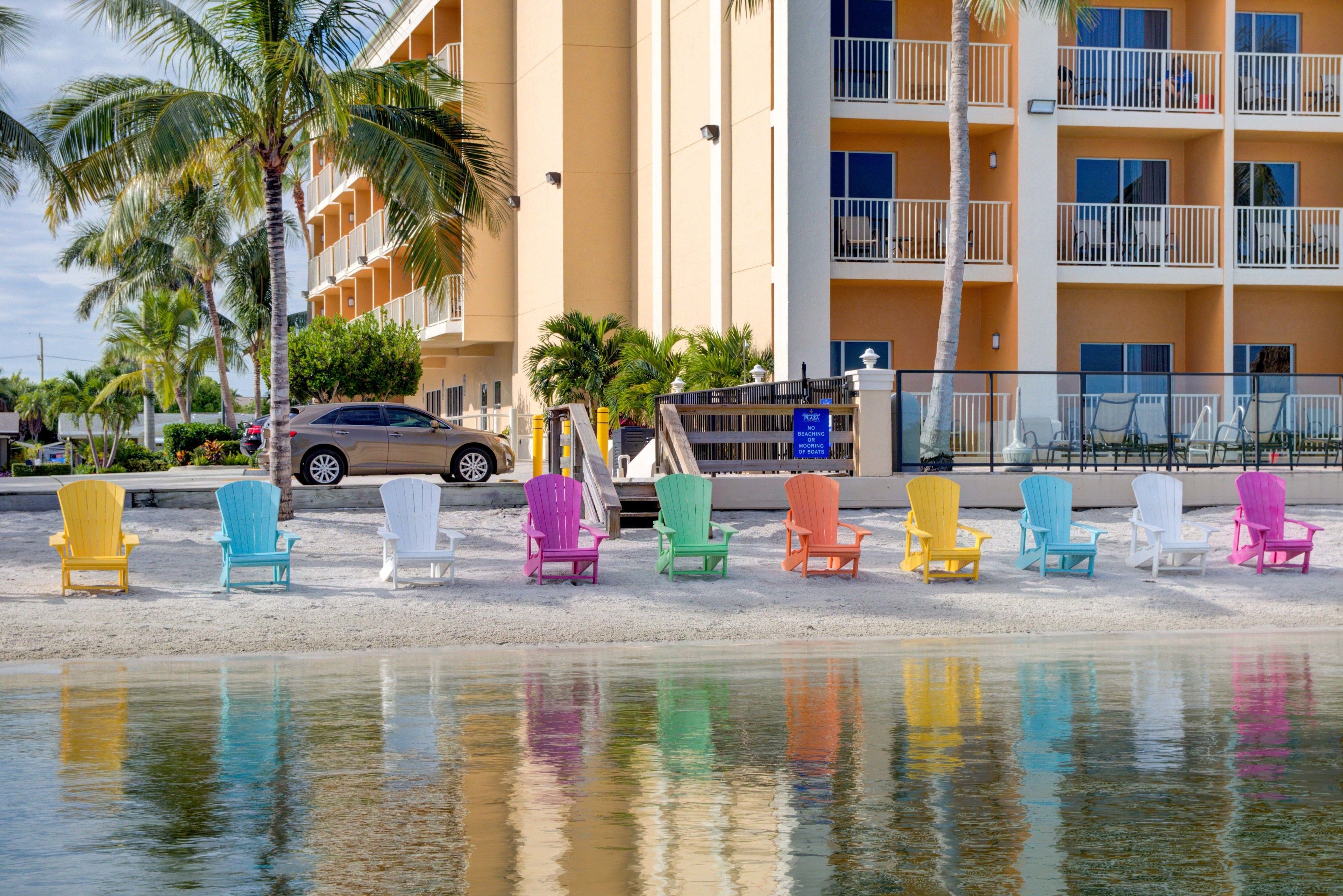 Um hotel na praia com cadeiras Adirondack coloridas alinhadas na areia, uma palmeira e um carro estacionado por perto.