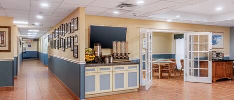 A hotel lobby with a reception desk, framed pictures on the wall, a kitchen area, and a dining table with chairs.