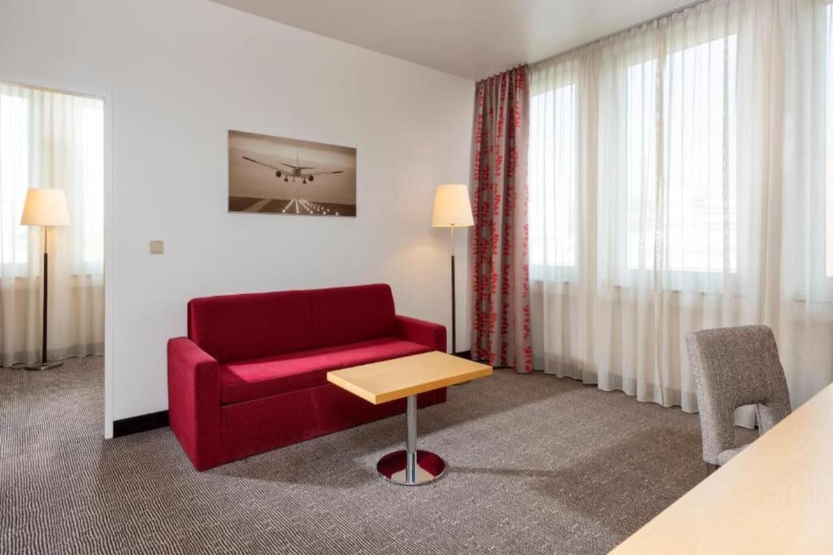 A hotel room with a red sofa, a small wooden table, a lamp, a chair, and a window with sheer curtains.