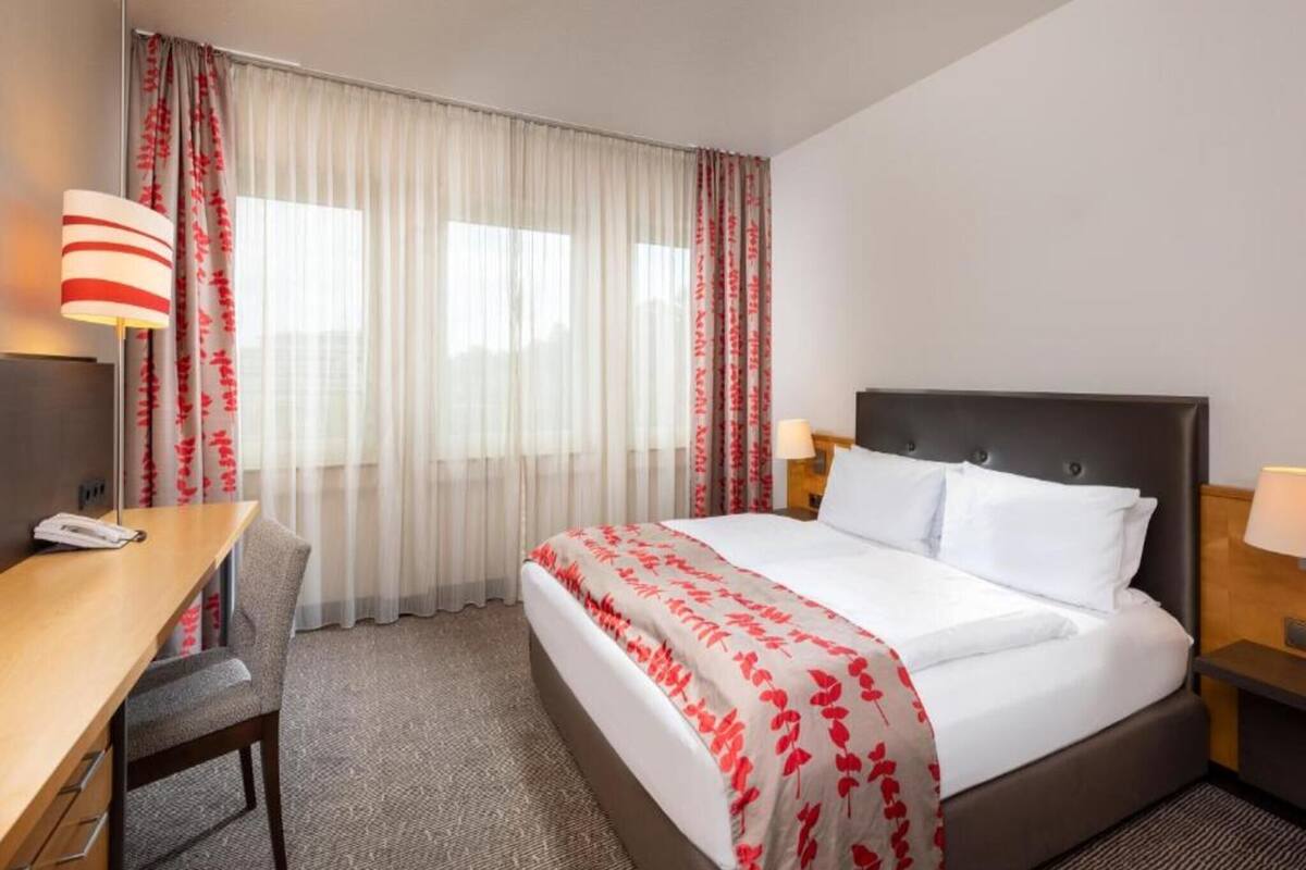 A hotel room with a bed, a desk, a chair, a nightstand, and a lamp.