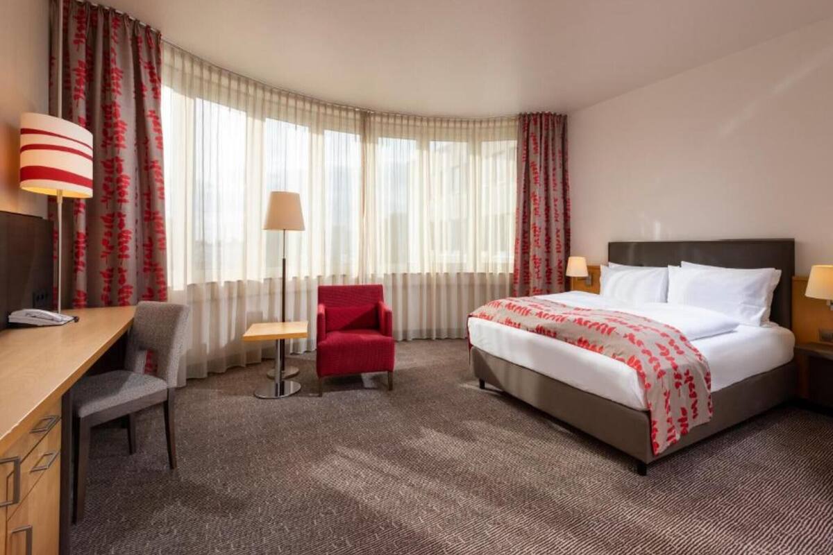 A hotel room with a bed, desk, chair, and a lamp.