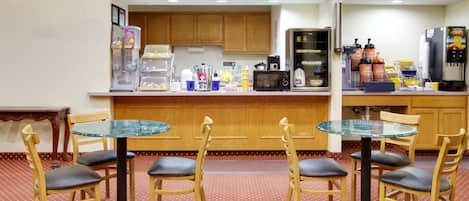 A small, cozy kitchen with wooden cabinets, a microwave, a coffee machine, and various food items. There is a round glass table with four chairs in front of the kitchen.