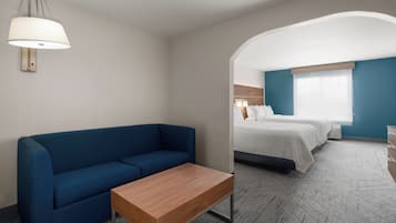 A hotel room with a blue sofa, a wooden side table, a bed with white bedding, and a large window.