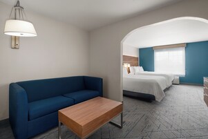 Habitación de hotel con un sofá azul, una mesita de madera, una cama con ropa de cama blanca y un ventanal grande.