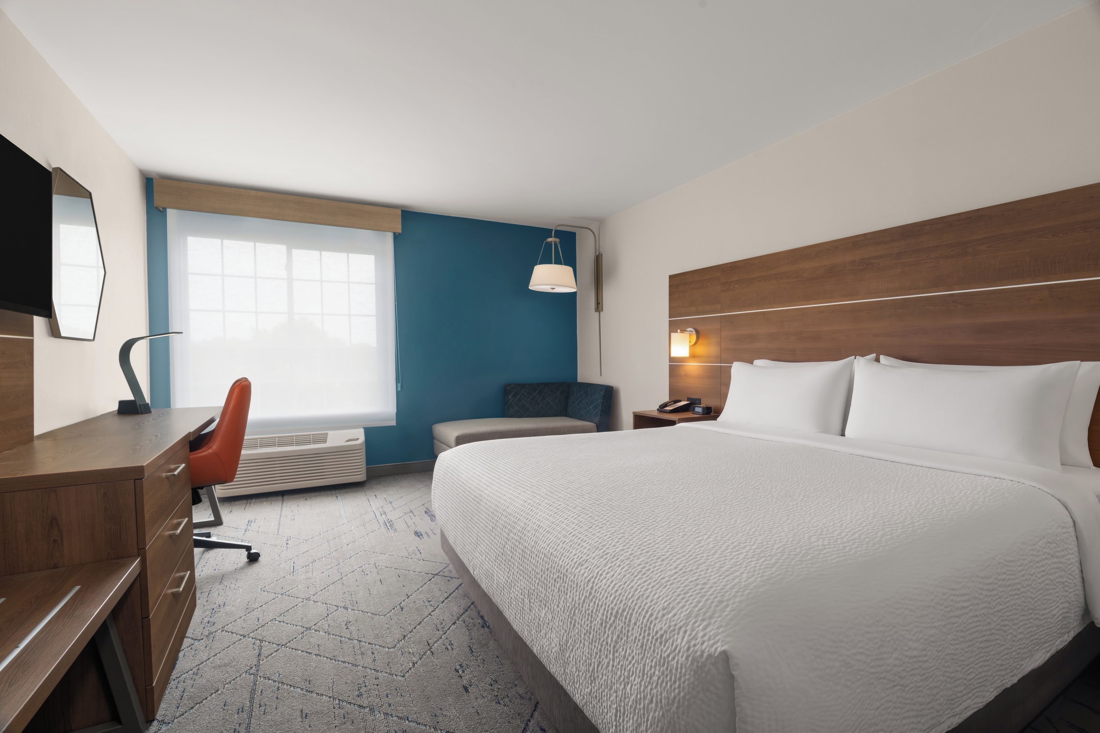Een hotelkamer met een groot bed, een bureau met stoel, een nachtkastje met lamp en een raam met jaloezieën.