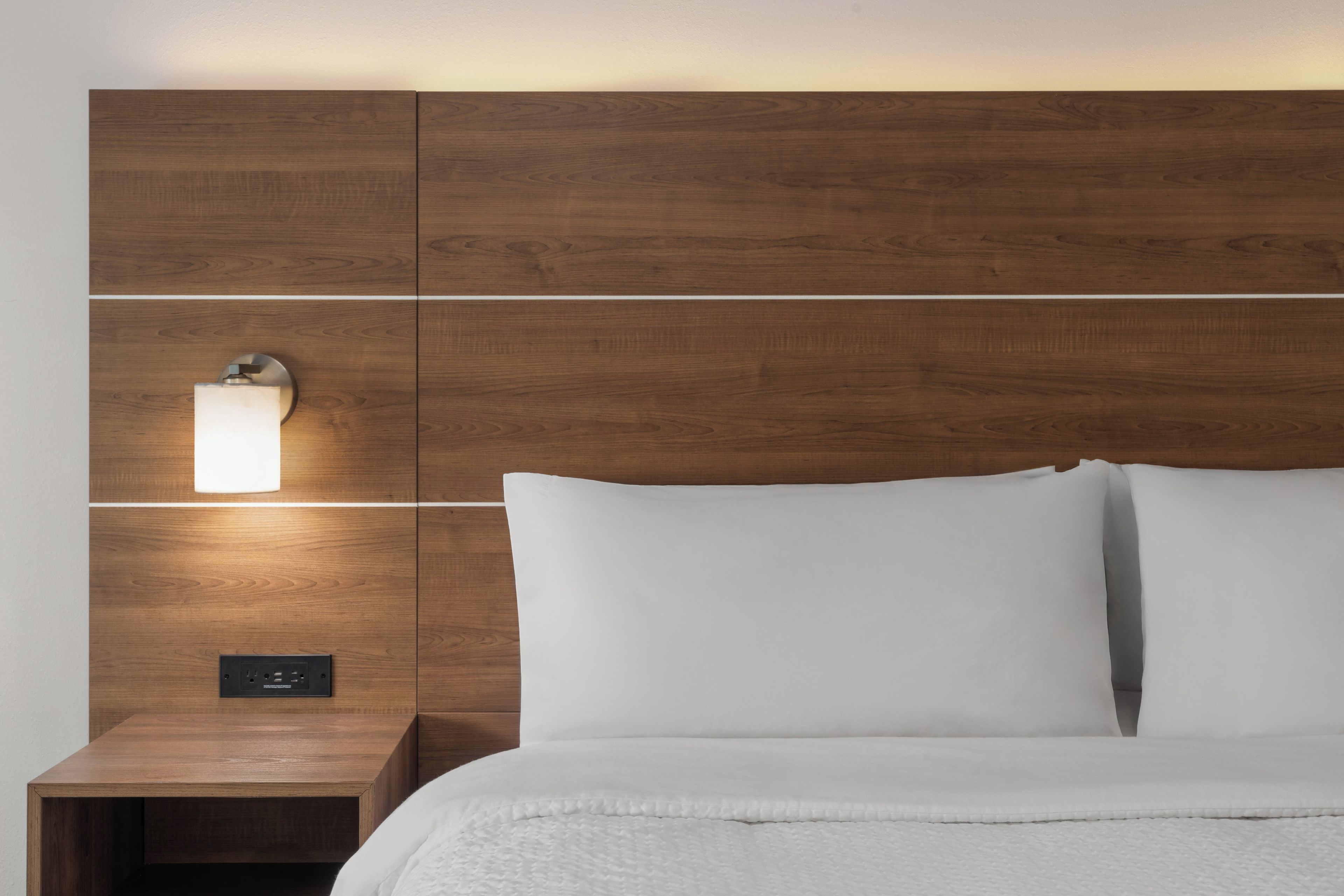 A neatly made bed with white linens and a wooden headboard with a wall-mounted light fixture.