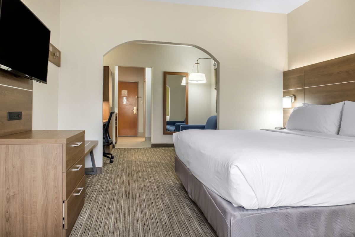 A hotel room with a large bed, a desk with drawers, a flat-screen TV, a chair, and a lamp.