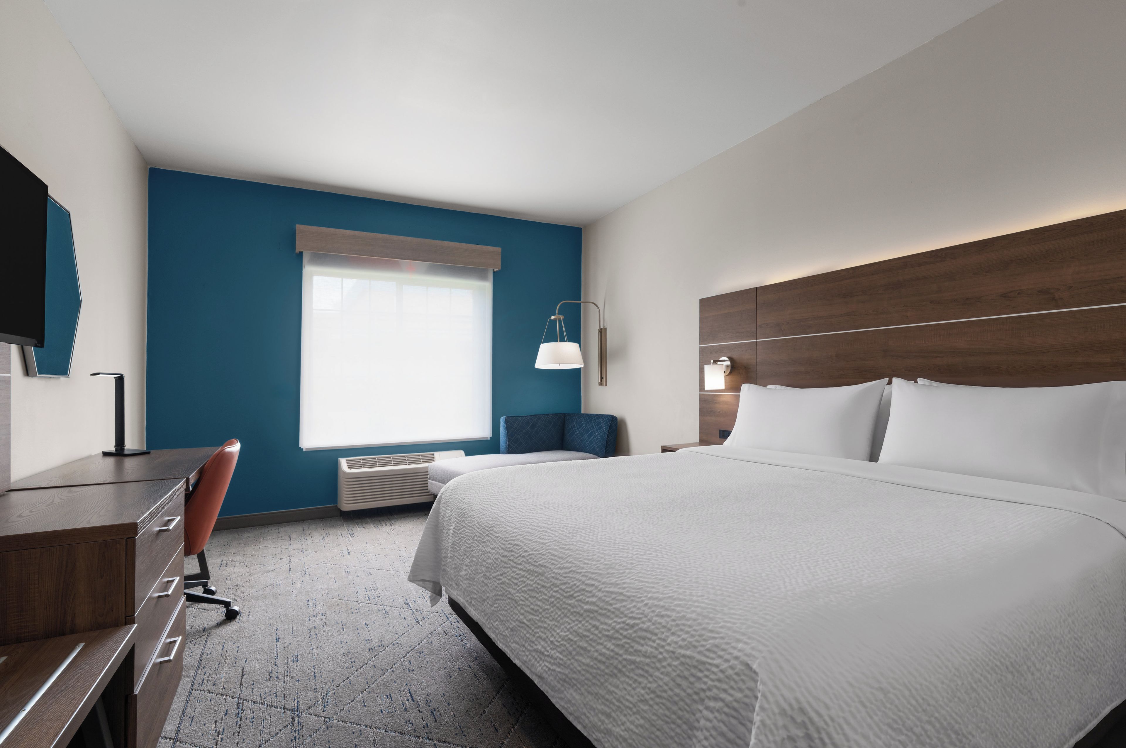 A hotel room with a large bed, a desk, a chair, a window with blinds, and a wall-mounted TV.