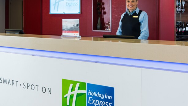 A Holiday Inn Express reception desk with a red wall, a bar area, and a sign indicating the hotel's name. - Holiday Inn Express Leeds City Centre by IHG (Leeds)