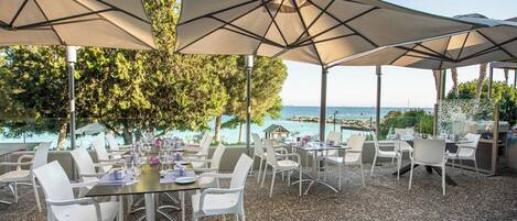 An outdoor dining area with tables and chairs, large umbrellas, and a view of the sea.
