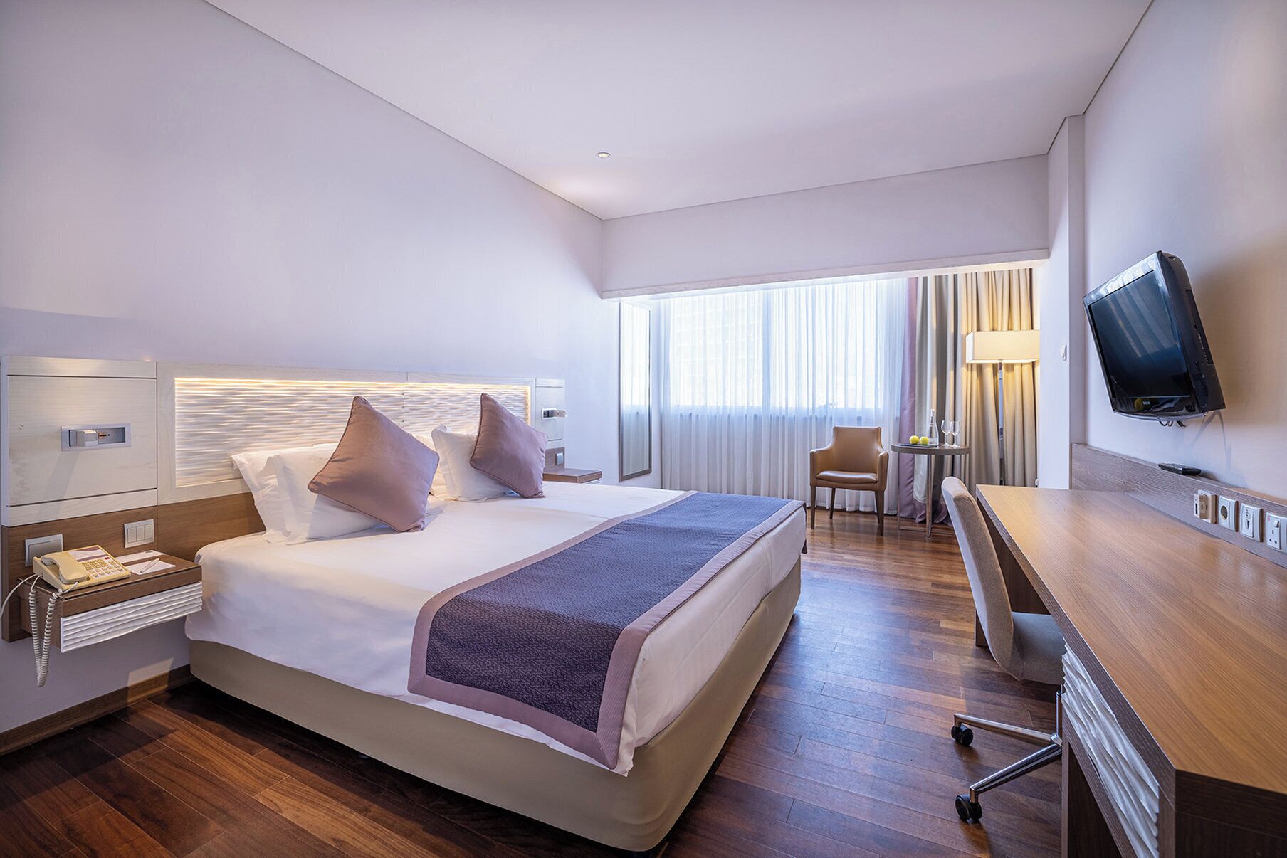 A hotel room with a large bed, bedside tables, a desk, a chair, a TV, and a window with curtains.