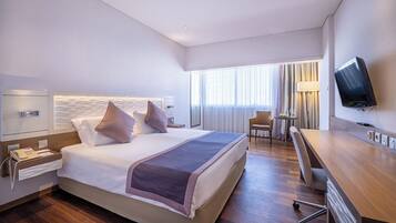 Habitación de hotel con una cama grande, mesitas de noche, un escritorio, una silla, un televisor y una ventana con cortinas.