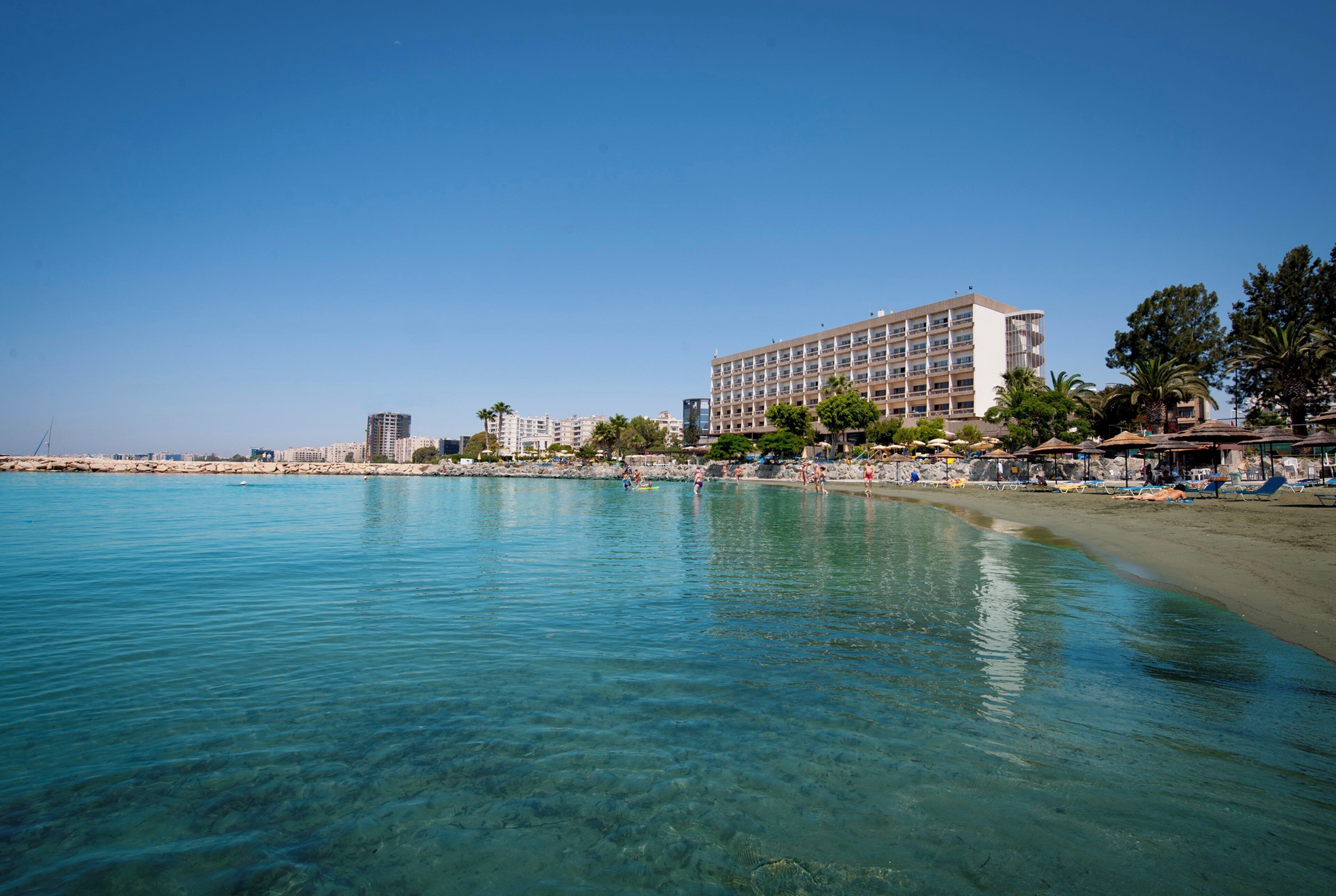 A beachfront hotel with clear blue waters, a sandy beach, and a row of palm trees.