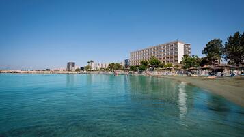 A beachfront hotel with clear blue waters, a sandy beach, and a row of palm trees.